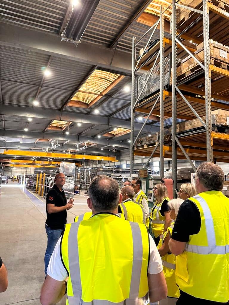 A group of people wearing yellow safety vests listen to a speaker in a large warehouse with rows of industrial shelving.