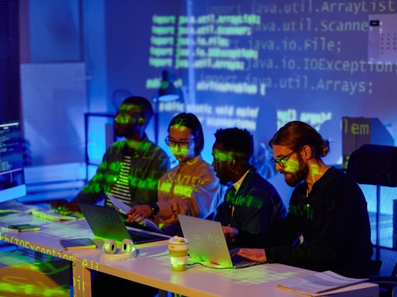 Four people sit at a desk working on laptops in a dim, blue-lit room with code projected onto them and the wall, suggesting a collaborative programming or hackathon environment.
