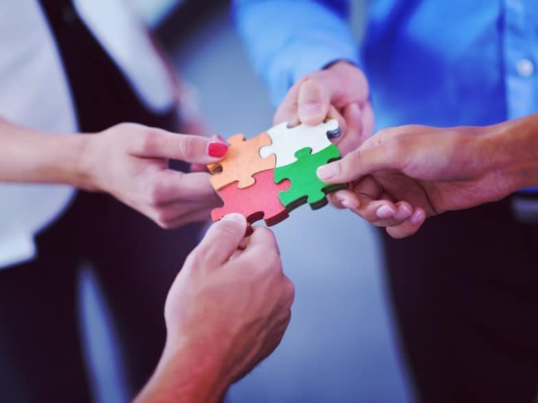 Four people each hold a puzzle piece, connecting them together to form a complete square, symbolizing teamwork and collaboration. The puzzle pieces are orange, white, green, and red.