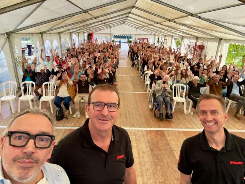 Three men in the foreground take a selfie in front of a large audience seated in a tent, all waving at the camera.