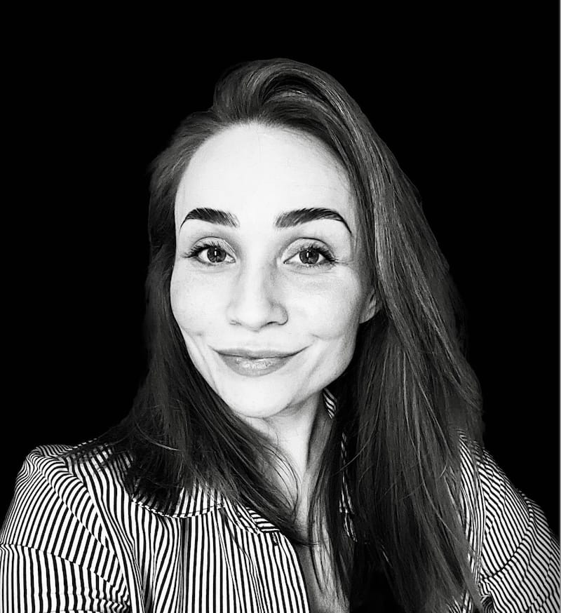 Black and white portrait of a woman with long hair, smiling softly. She wears a striped shirt and looks directly at the camera, against a solid black background.