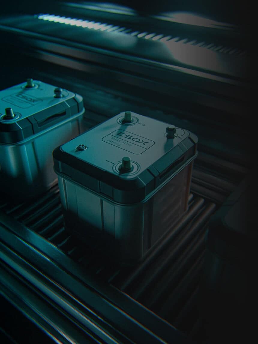 A close-up of a large, rectangular battery with metallic terminals on top, lit by blue-green lights, placed on a metal surface with other similar batteries in the background.
