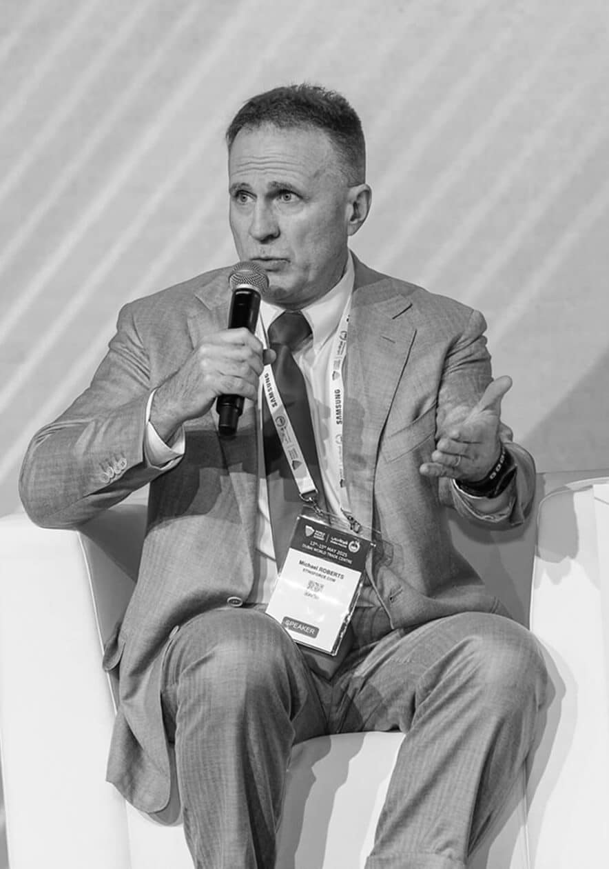 A man in a suit sits on a white chair, speaking into a microphone. He gestures with his left hand, wearing a conference badge around his neck. The image is in black and white.
