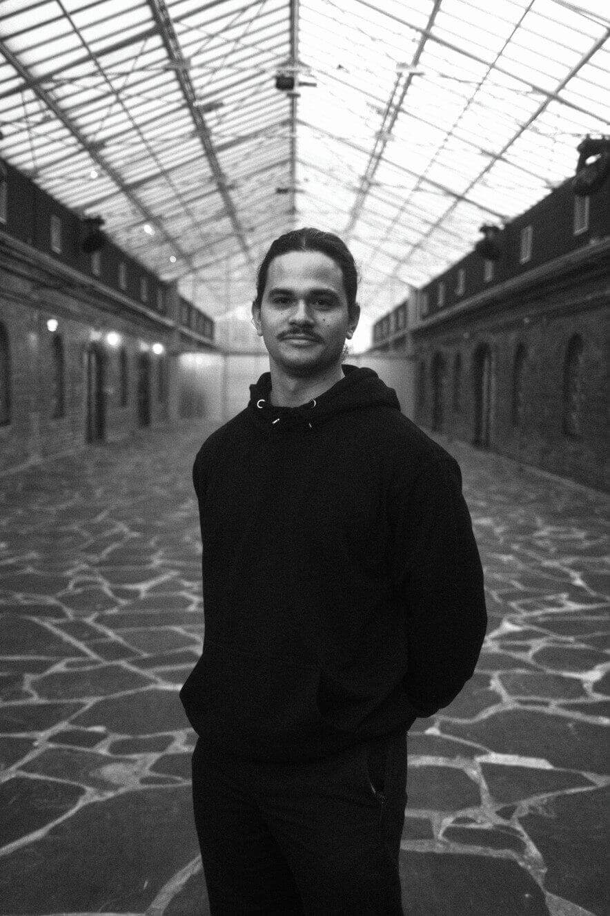 A person wearing a dark hoodie stands in the center of a large corridor with a glass ceiling and brick walls, looking at the camera. The space has a patterned stone floor and ambient lighting.