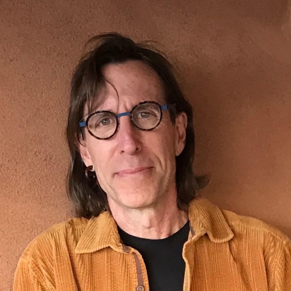 A man with glasses and a jacket standing in front of a tan wall.