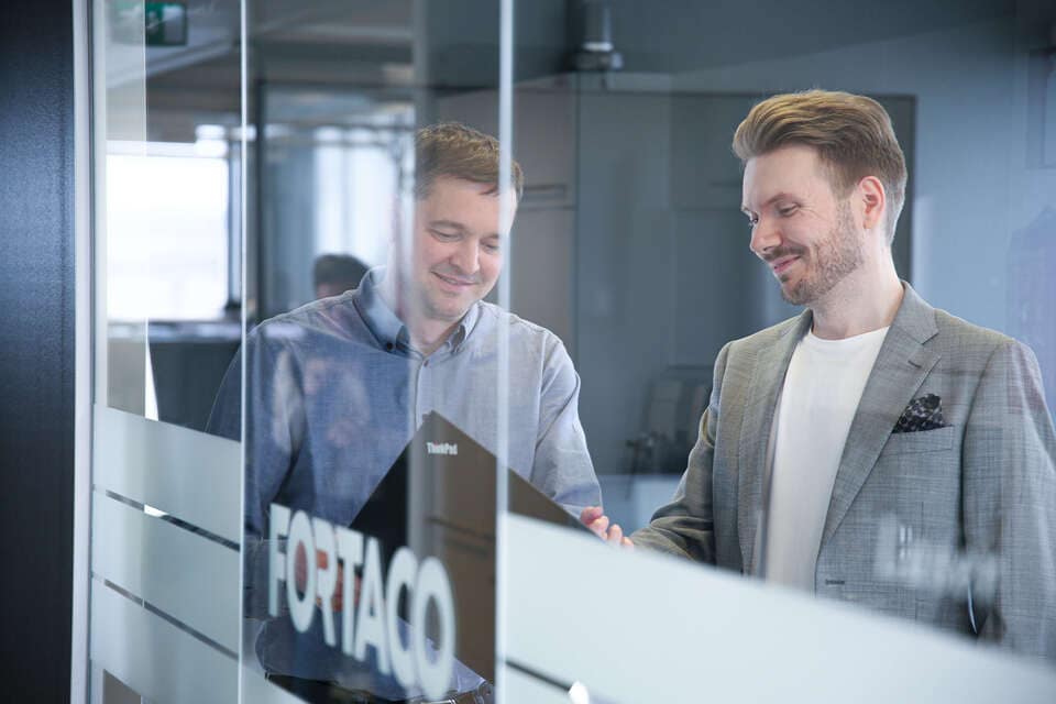 Two men in business attire stand in an office behind a glass wall with 'FORTACO' written on it, smiling and looking at a laptop together.