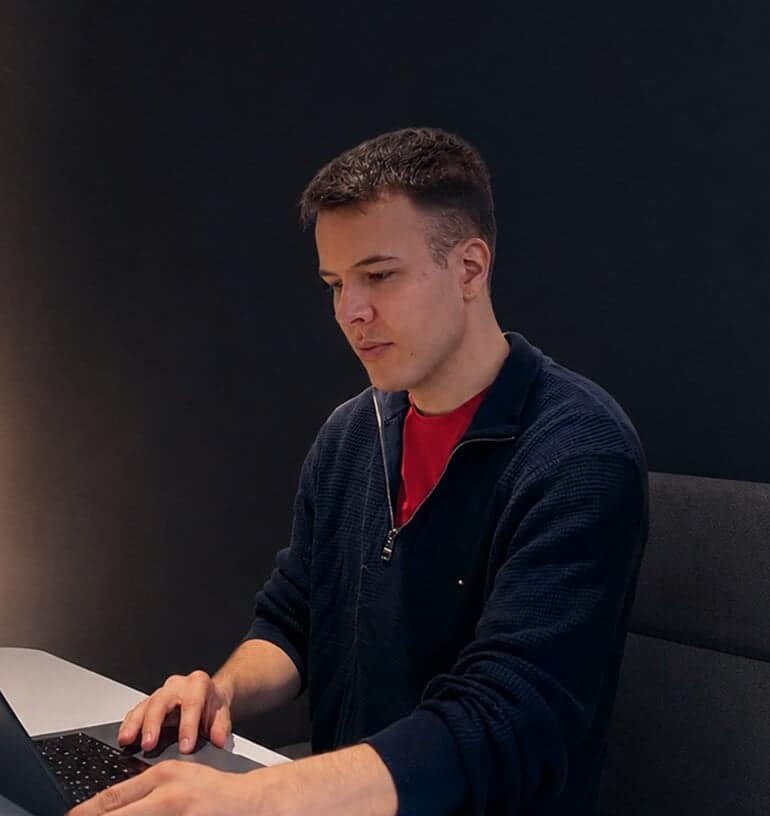 A person with short brown hair sits at a desk, typing on a laptop. They wear a dark zip-up jacket over a red shirt, with a dark background behind them.